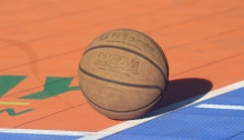 Image shows a basketball lying on a court during a bright, sunny day. The basketball is orange and slightly worn. It is resting at the right corner of the free-throw line, which is painted white. Part of the court is bright orange, but a strip of blue is featured to the right and directly in front of the ball. The ball casts a shadow onto the court.