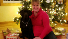 Image shows Janell Groskreutz, right, with her guide dog, Sully, a black lab. Janell and Sully are sitting close to each other on the floor and Janell has a bright, beautiful smile on her face. She is pleased to be finished with holiday shopping, eagerly anticipating Christmas Eve when her family opens their presents and she hears excitement and happiness in their reactions. A lighted, fully-decorated Christmas tree sparkles in the background and presents are spread along the hardwood floor, surrounding Sully and Janell.